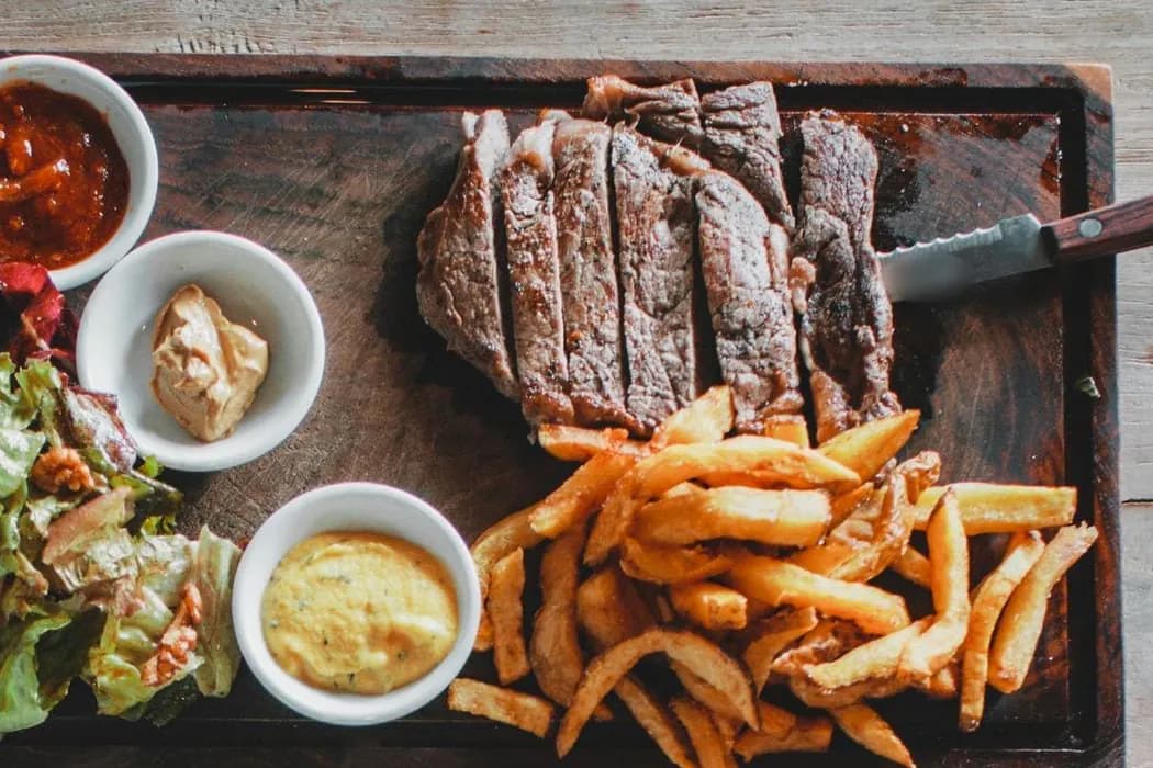Tábua com bife fatiado, batatas fritas, salada verde e molhos em mesa de restaurante